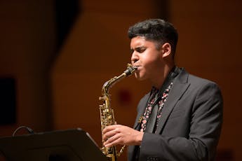 A student performer dressed in a suit jacket and with a colorful dress shirt plays a saxophone at a podium.