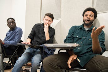 Three students sitting at desks along a white wall participate in a discussion during an Arts Management and Entrepreneurship class.