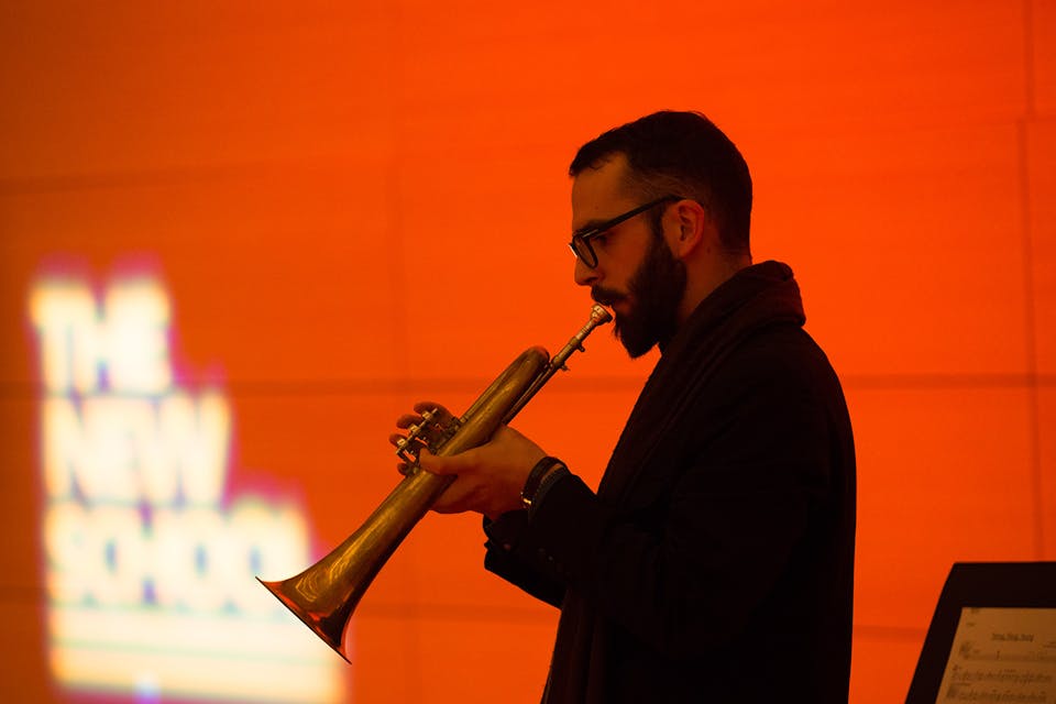 "A student performer dressed in black plays the trumpet in front of a red-orange wall with the words “The New School” illuminated in the background