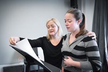 Vocalist reads music on podium with a faculty member supporting their posture. The vocalist holds their diaphragm for proper breathing.