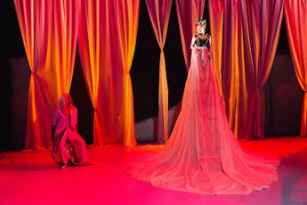 A red-light flooded stage: a towering performer in elaborate dress and regal jewelry gazes down on another, who appears startled.