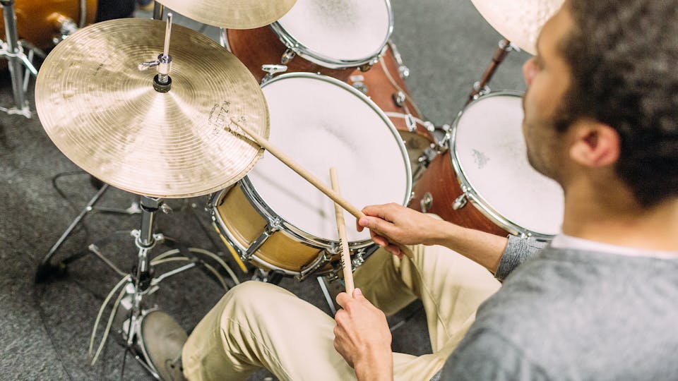 A student in a gray shirt and khaki pants plays a drum set.