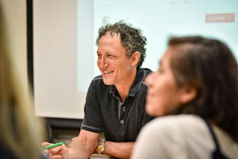 A professor smiles at students in a classroom with a dropdown screen behind.