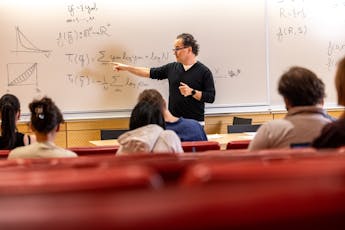 A profesor points to a formula on a white board in front of a roomful of students.