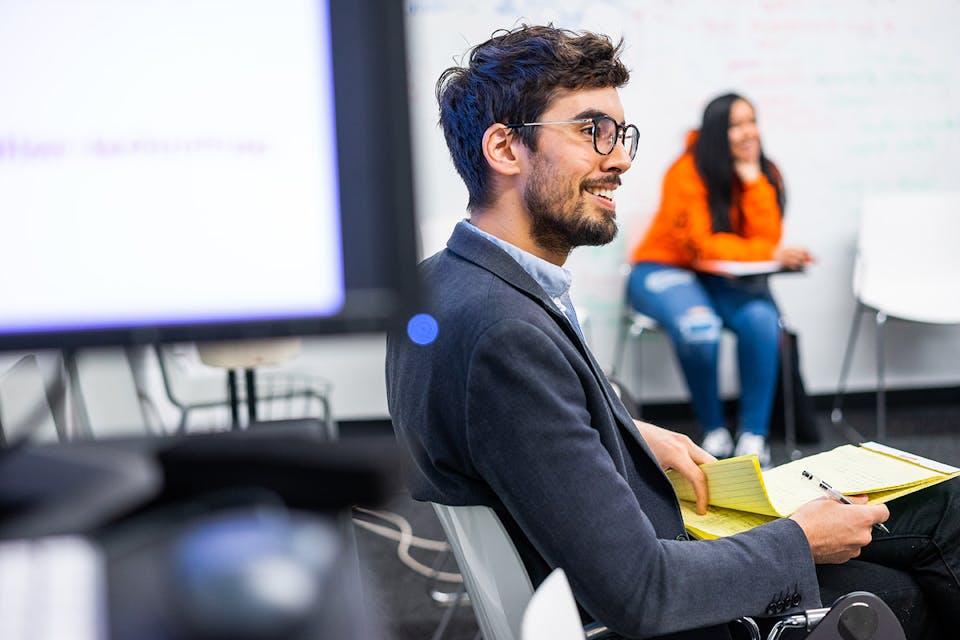 A person seated in a classroom near a projection screen smiles while looking to the side and paging through a notepad.