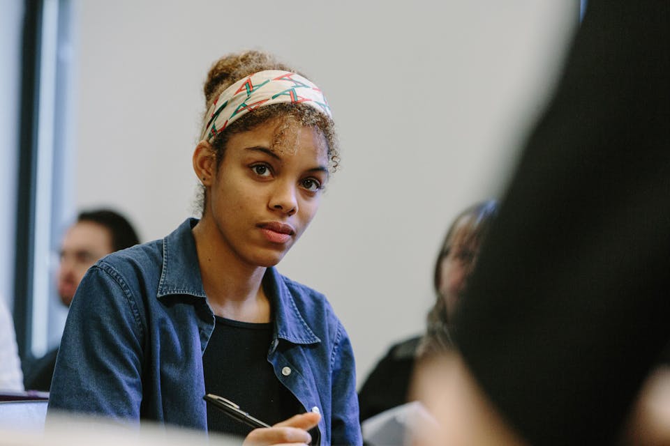 A seated student in a jean jacket and colorful headband looks intently with pen in hand in a classroom.