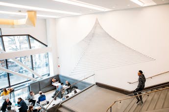 Overhead view of students at tables in a study space with a pyramid graphic on the wall. Student descends a staircase in the foreground.