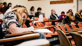 In a lecture hall, a student takes notes while classmates appear in the background. The space is furnished with orange chairs.