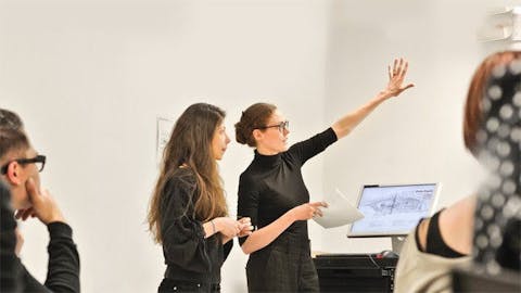 A standing professor demonstrates something with their arm up. A student stands next to her; others are seated. Everyone is wearing black.